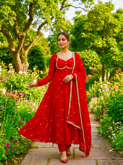 Woman in a red traditional outfit standing in a garden with greenery and flowers.